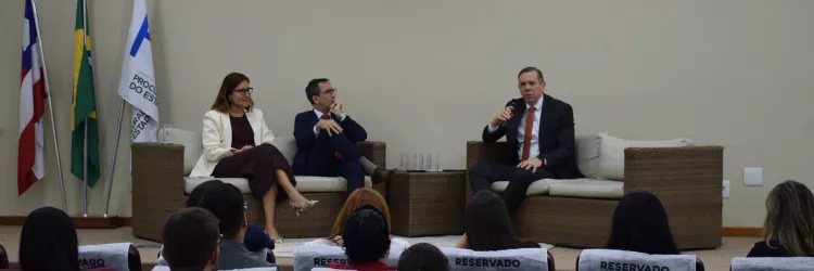 Três pessoas estão sentadas em poltronas no palco, participando de uma conversa. Uma mulher veste blazer claro e vestido vinho. Ao lado dela, um homem de terno azul e gravata vermelha. Outro homem, de terno escuro e gravata laranja, segura um microfone e fala. Atrás deles, há quatro bandeiras: da Bahia, do Brasil, da PGE-BA e uma quarta não identificada. À frente, o público assiste atento.