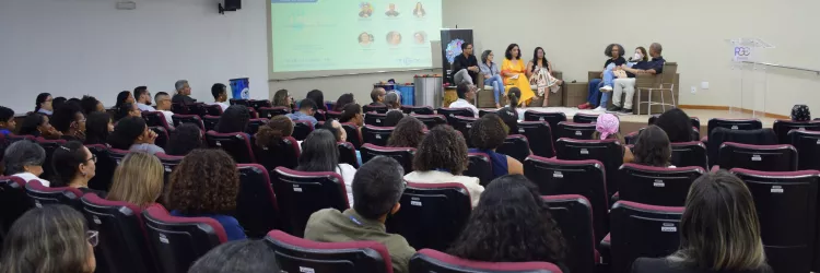 A imagem mostra um auditório com várias pessoas sentadas em cadeiras pretas de encosto alto, voltadas para a frente. No palco, à direita, há um grupo de oito pessoas sentadas em poltronas, participando de uma roda de conversa. Uma tela de projeção ao fundo exibe informações do evento, com fotos e nomes de participantes. À esquerda, o público observa atentamente. À direita, há um púlpito transparente e uma pessoa em pé, interpretando em Libras. O ambiente é iluminado por luzes de teto embutidas.