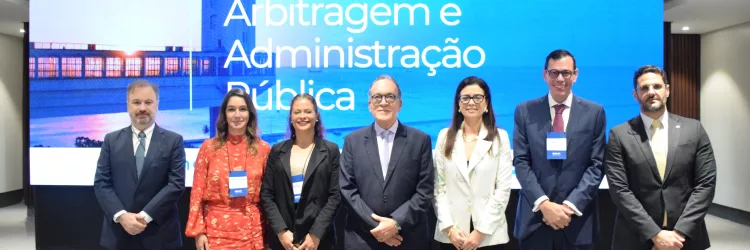 Sete pessoas estão em pé, lado a lado, sorrindo para a foto, em frente a uma grande tela com os dizeres “Arbitragem e Administração Pública”. Quatro homens de terno e gravata e três mulheres bem vestidas, sendo uma com terno floral claro, uma com roupa preta e outra com conjunto vermelho estampado. Todos usam crachá azul pendurado no pescoço. O ambiente é fechado, bem iluminado e com aparência profissional.