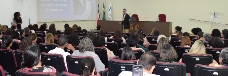 Auditório cheio de pessoas sentadas em poltronas vermelhas. À frente, um homem de blazer preto fala com microfone, enquanto no telão aparece a frase: “Afinal, não quero que ache um tédio a palestra”. Ao fundo estão as bandeiras do Brasil, da Bahia e outra institucional.