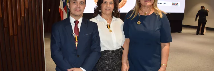 Três pessoas posam para foto em frente ao mesmo telão da homenagem. Um homem de terno azul, uma mulher de blusa branca e saia escura, e outra de vestido azul. Todos usam medalhas douradas e sorriem para a câmera.