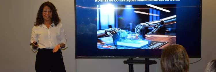 Palestrante apresenta o tema “O Código da Lei nº 14.634/2023: A Chave de Leitura das Normas de Contratações Administrativas na Bahia”. Ela está de pé ao lado de uma tela que exibe uma imagem ilustrativa com braços robóticos manuseando dados digitais, indicando abordagem tecnológica no contexto jurídico. O público acompanha sentado, atento à exposição.