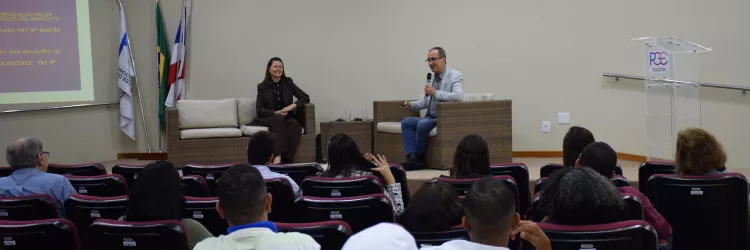 A imagem mostra uma palestra em andamento em um auditório. À frente, um homem fala ao microfone, sentado em uma poltrona de vime com almofadas claras, enquanto uma mulher, também sentada em poltrona semelhante, o observa sorridente. À esquerda, há um telão com apresentação projetada e bandeiras posicionadas ao lado. O público, composto por homens e mulheres, está sentado em cadeiras vermelhas voltadas para o palco. O ambiente é iluminado e organizado.