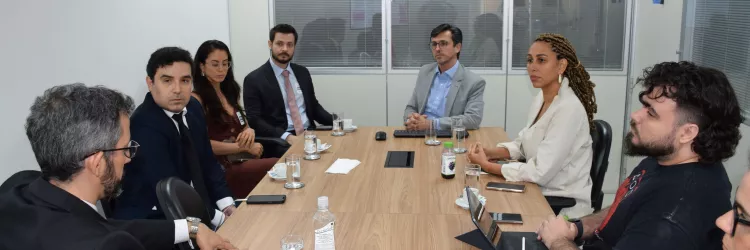Reunião em uma sala com oito pessoas sentadas ao redor de uma mesa retangular de madeira. Elas estão em conversa atenta, com copos de água, canecas, garrafas e cadernos sobre a mesa. O ambiente é corporativo, com paredes brancas, janelas de vidro e iluminação clara. As pessoas usam roupas formais e demonstram concentração no diálogo.