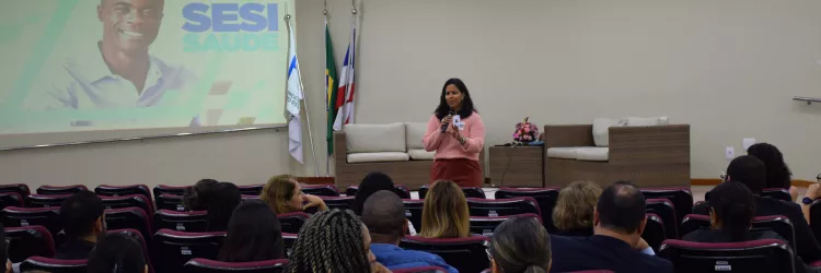 Em um auditório, uma mulher fala ao público segurando um microfone. Ela veste blusa rosa e calça marrom. À frente, há várias fileiras de cadeiras ocupadas por pessoas atentas. No fundo, há um telão com a imagem de um homem sorridente e o texto “SESI SAÚDE”. Próximo ao telão, estão três bandeiras e dois sofás de fibra com almofadas claras. O ambiente é bem iluminado e organizado, com clima de palestra ou apresentação.