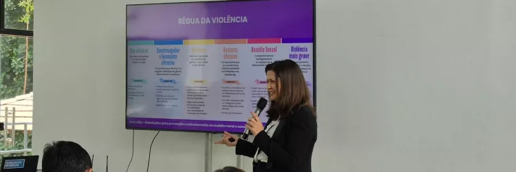 Uma mulher está em pé, segurando um microfone e apresentando em frente a uma tela grande. Ela tem cabelos castanhos, usa blazer preto e blusa clara. Na tela aparece o título “Régua da Violência”, com uma tabela colorida em cinco colunas, que classificam diferentes níveis de violência, de “Não ofensivo” até “Violência mais grave”. Algumas pessoas estão sentadas assistindo à apresentação, em um ambiente claro, com paredes brancas e janelas amplas.