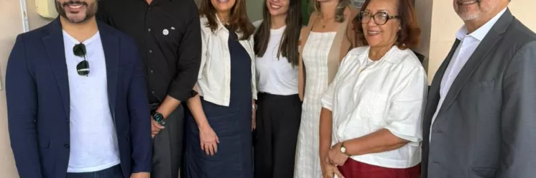 Sete pessoas estão em pé, sorrindo para a câmera, em frente a uma parede bege com uma porta de vidro e uma faixa com o logo "PGE". São quatro mulheres e três homens, vestidos com roupas sociais ou casuais elegantes. À esquerda, um homem de blazer azul e camiseta branca. Ao centro, duas mulheres usam roupas claras. À direita, uma senhora com blusa branca e calça vermelha segura uma bolsa clara, ao lado de um homem de terno cinza. Todos parecem felizes e em um ambiente profissional.