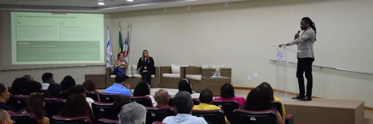 Auditório com pessoas sentadas em poltronas bordô assistindo a uma palestra. Um homem em pé, de blazer claro e calça escura, fala ao microfone em um púlpito transparente com o logotipo da PGE. No fundo, há duas mulheres sentadas em poltronas próximas a bandeiras do Brasil, da Bahia e da PGE-BA. Um telão projeta o tema “CPAD e Assessoria Jurídica na Gestão Documental”. O ambiente é bem iluminado, com teto branco e luzes embutidas.