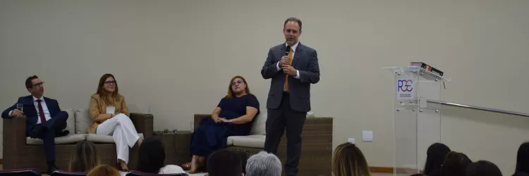 Auditório com várias pessoas sentadas em cadeiras estofadas vinho, voltadas para o palco. No palco, três pessoas estão acomodadas em sofás de fibra: um homem de terno azul, uma mulher de blazer bege e calça branca e outra de vestido azul-marinho. Em pé, à direita, um homem de terno cinza e gravata amarela fala ao microfone próximo a um púlpito transparente com o logotipo da PGE-BA. O fundo é uma parede bege lisa e o ambiente é iluminado por luzes embutidas no teto.