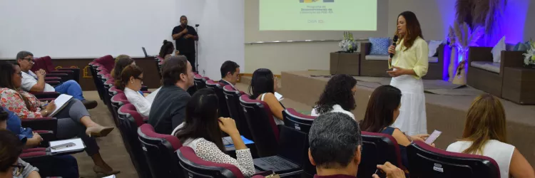 Uma mulher fala em pé diante de um auditório, segurando um microfone. Ela veste blusa amarela clara e saia branca. As pessoas estão sentadas em cadeiras vinho, observando e fazendo anotações. No fundo, há um telão com o logotipo do “Programa de Desenvolvimento de Lideranças da PGE-BA”. Um homem ao lado do auditório faz interpretação em Libras. O ambiente é iluminado e organizado, com clima de evento institucional.