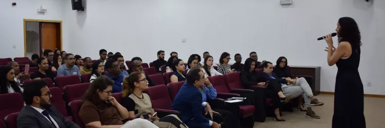 A imagem mostra uma mulher de vestido preto falando ao microfone para um público sentado, atento. As pessoas estão bem-vestidas, em roupas formais ou sociais, e o ambiente tem cadeiras estofadas vinho, paredes brancas e iluminação clara. A maioria dos participantes parece jovem e há diversidade de gênero e etnia entre os presentes.