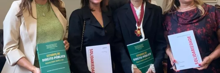 Quatro mulheres estão em pé, sorrindo, lado a lado, em um ambiente formal com quadros nas paredes. Duas delas seguram um livro verde intitulado "Estudos de Direito Público" e as outras duas seguram um livro branco com a palavra "Solidariedade" em vermelho. A terceira mulher, de cabelos grisalhos e roupa preta, usa uma medalha no pescoço. Todas estão bem-vestidas e aparentam estar em um evento jurídico-acadêmico.