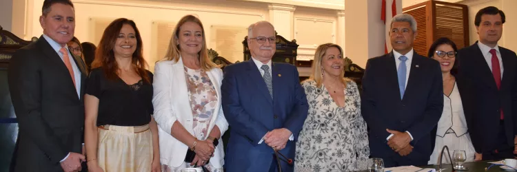Oito pessoas posam em pé atrás de uma mesa de madeira verde, com microfones e copos d’água sobre ela. Todas estão sorrindo e vestidas de forma formal, com ternos, vestidos e blazers. O ambiente é iluminado e tem bandeiras da Bahia e do Brasil ao fundo, além de detalhes clássicos nas paredes e no teto, indicando um evento institucional.