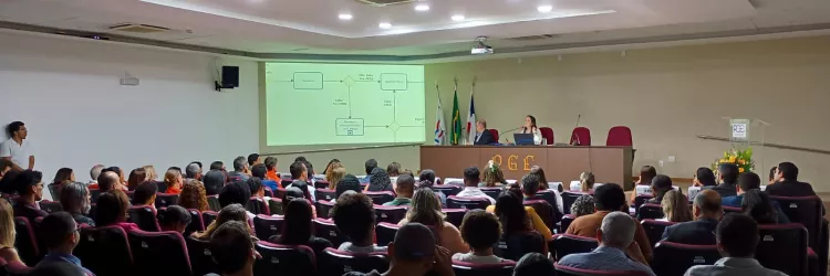 Visão geral de um auditório com uma plateia sentada em poltronas vermelhas escuras, voltada para um palco. A sala é iluminada e tem um teto rebaixado.  No palco, uma mesa escura com duas pessoas, com um pódio de acrílico à direita. Atrás delas, três bandeiras e uma tela de projeção exibem um fluxograma simples e claro. A plateia é diversificada e preenche o auditório.