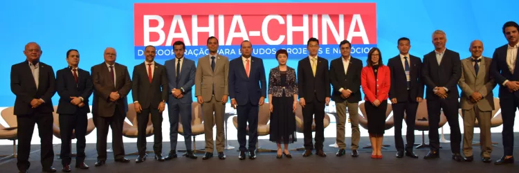 A imagem mostra quinze pessoas em um palco, sendo treze homens e duas mulheres, todas vestidas com roupas formais. Elas estão alinhadas, olhando para a câmera e sorrindo. Ao fundo, há uma grande tela azul com um banner vermelho onde se lê “III FÓRUM BAHIA-CHINA”. O ambiente é bem iluminado e o palco tem um tapete cinza escuro.