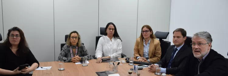 Seis pessoas estão sentadas ao redor de uma mesa de madeira em uma sala de reunião. Três mulheres estão de um lado, e uma mulher e dois homens do outro, totalizando seis pessoas. Elas olham atentamente para frente, com expressões sérias e concentradas. Sobre a mesa há copos com água, xícaras de café, celulares, cadernos e canetas. O ambiente é claro, com paredes brancas e iluminação de teto.