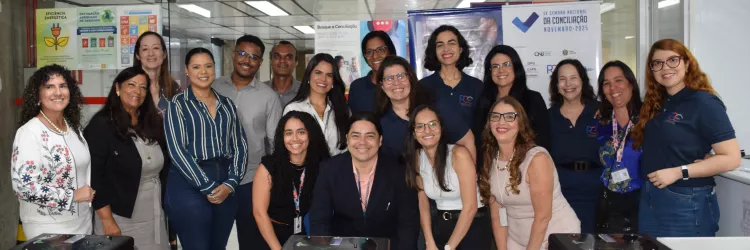 Um grupo de vinte pessoas, composto por homens e mulheres, posa sorrindo para a câmera em um ambiente interno. A maioria veste roupas sociais ou casuais escuras. O grupo está de pé, exceto por um homem no centro, agachado, e uma mulher à sua frente. Eles se agrupam próximos a duas catracas pretas, com a palavra "ENTRADA" em branco. Ao fundo, painéis e cartazes coloridos estão em exposição. As luzes de teto são fluorescentes.