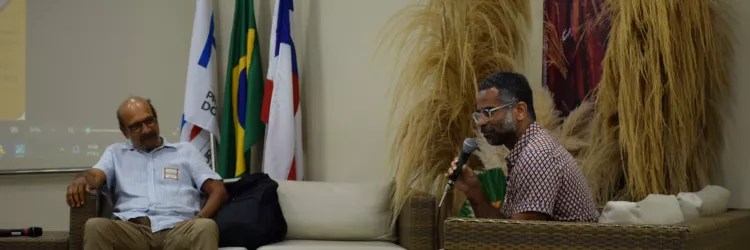 Dois homens sentados nas poltronas de fibra conversam. Um deles segura o microfone, enquanto o outro escuta com expressão atenta, com o braço apoiado na poltrona. O cenário tem bandeiras ao fundo, arranjos de folhas secas e o painel com a imagem da mulher negra. O clima é de diálogo durante um debate.