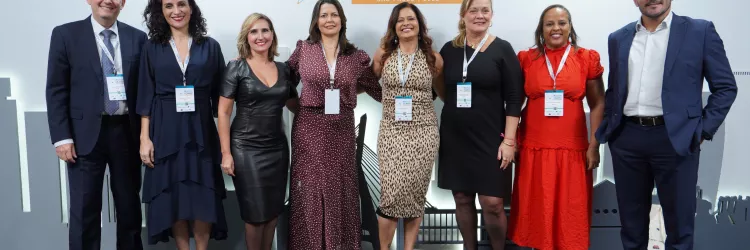  A imagem mostra oito pessoas posando para foto em frente a um painel de evento. Elas estão alinhadas lado a lado, sorrindo, e usam roupas sociais e crachás. Há homens e mulheres no grupo, todos em postura formal e amistosa. O painel atrás é claro, com logotipos repetidos. O ambiente parece ser de congresso ou encontro profissional.