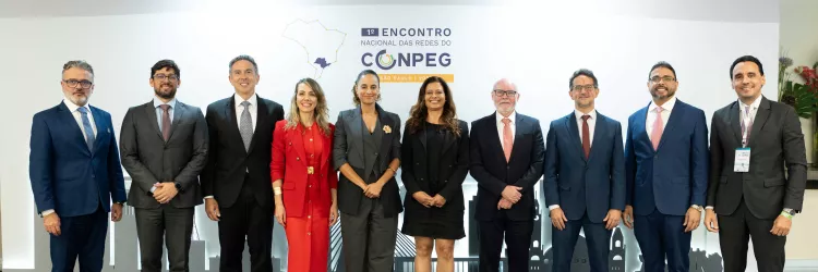 A imagem mostra um grupo de dez pessoas alinhadas lado a lado, posando para a foto em um evento. São sete homens e três mulheres, todos vestidos de forma formal, com ternos ou vestidos sociais. Eles estão em frente a um painel branco com o logotipo do “1º Encontro Nacional das Redes do CONPEG – São Paulo”. O chão é de cor neutra e o ambiente é bem iluminado. Todos sorrirem levemente, aparentando profissionalismo e cordialidade.