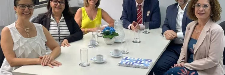Seis pessoas estão sentadas em volta de uma mesa de reuniões branca, em um ambiente de escritório claro e organizado. Três mulheres estão de um lado e uma mulher e dois homem do outro. Todos sorriem e estão vestidos de forma social. Sobre a mesa há xícaras de café, copos de água, uma jarra, uma revista e um pequeno vaso com flores azuis. Ao fundo, há uma janela grande com vista para árvores e um quadro com a imagem de um barco à vela.