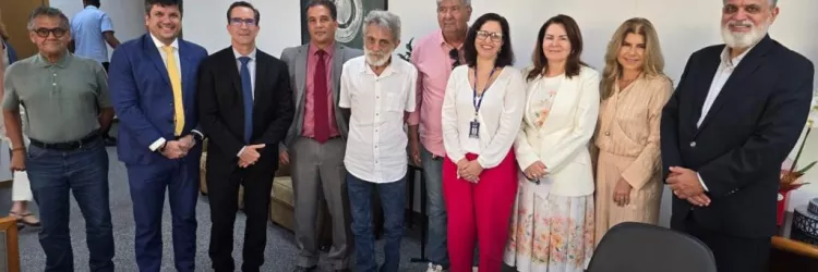 A imagem mostra um grupo de dez pessoas em pé, posando para uma foto em um ambiente de escritório. São sete homens e três mulheres. Alguns usam ternos e roupas formais, enquanto outros estão vestidos de forma mais casual. Ao fundo, há uma parede branca, um quadro, uma porta de madeira e móveis de escritório. À frente, há uma mesa com copos e papéis. Todos olham para a câmera e sorriem.