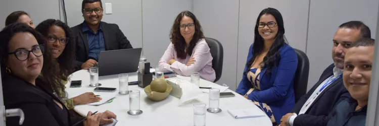 A foto mostra um grupo de pessoas sentado ao redor de uma mesa redonda branca em uma sala de reunião. Há copos de água, celulares e cadernos sobre a mesa. Todos observam alguém fora do quadro, alguns sorrindo. Ao fundo, há uma TV desligada na parede. O ambiente é claro e organizado.