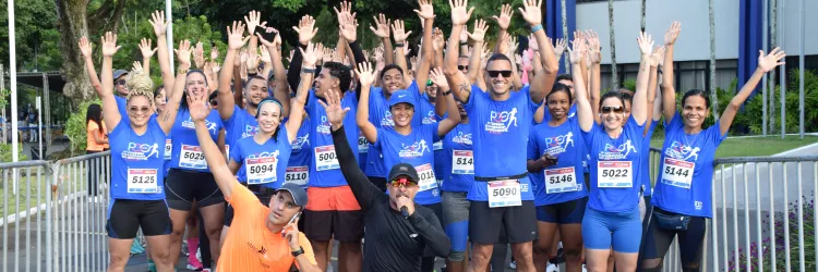 Foto que mostra um grupo de cerca de 20 corredores de rua, sorridentes, de camiseta azul com o logo da corrida. Eles estão em pé, com os braços levantados, logo após a linha de chegada de uma corrida. Dois homens estão agachados na frente.