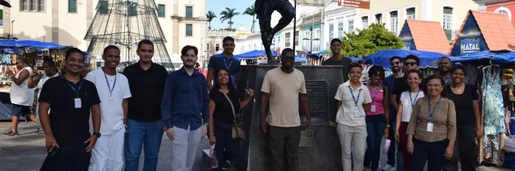 Um grupo grande posa para foto em frente à estátua de Zumbi dos Palmares, em uma praça com prédios históricos ao fundo. Todos estão alinhados ao redor do monumento, sorrindo. Há barracas de artesanato e uma estrutura de árvore de Natal próxima. O céu está azul e a luz do dia ilumina claramente o local.