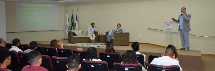 A imagem mostra um auditório onde um homem está em pé, falando em um púlpito à frente de uma plateia. Ele veste paletó cinza e segura um microfone. Ao fundo, há um telão verde com texto amarelo sobre "Principais doenças masculinas", listando várias condições de saúde. Três bandeiras estão ao lado do púlpito. Na frente do palco, dois homens estão sentados em um sofá. O público está sentado em cadeiras vermelhas e pretas, assistindo à apresentação. O ambiente é claro e formal, com iluminação no teto.