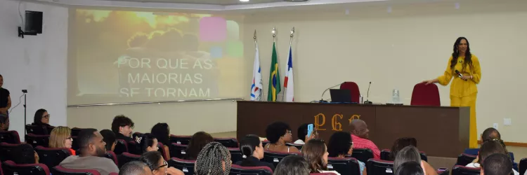 Auditório cheio, com pessoas sentadas em cadeiras acolchoadas, assistindo a uma palestra. No palco, uma mulher de roupa amarela fala ao público ao lado de uma mesa com computador e microfone. Ao fundo, um telão mostra um slide com a frase “Por que as maiorias se tornam…”. Há bandeiras posicionadas atrás da mesa e iluminação de teto ligada.