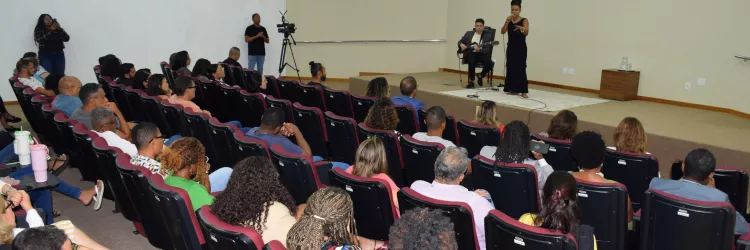 A plateia observa a apresentação no palco, onde a cantora e o músico realizam a performance. A imagem mostra a visão geral da sala, com todas as fileiras ocupadas