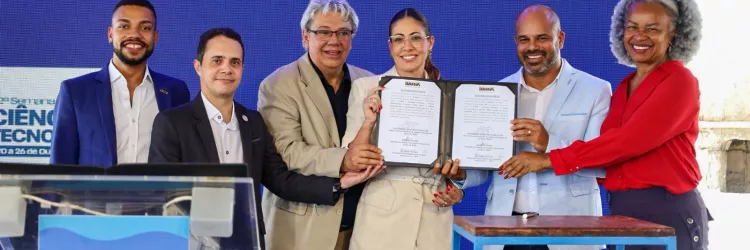 Seis pessoas, quatro homens e duas mulheres, estão em pé em frente a um painel azul. O grupo segura um documento em conjunto.  Da esquerda para a direita: um homem negro de blazer azul; um homem branco de camisa social preta; um homem branco de blazer bege; uma mulher branca de blazer e calça bege claro; um homem negro de blazer azul claro; e uma mulher negra de camisa vermelha. Todos sorriem.