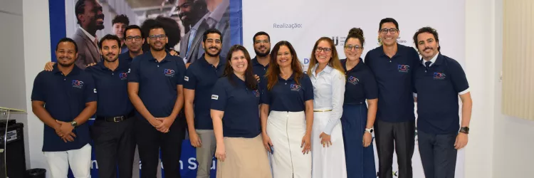 Um grupo de doze pessoas posa sorridente em um ambiente interno, diante de um grande banner azul e branco com o texto “XX Semana Nacional da Conciliação – Novembro 2025”. A maioria veste camisa polo azul-marinho com o logotipo da PGE, calças sociais ou saias em tons neutros. O grupo está lado a lado, demonstrando integração e alegria. O local tem piso claro, parede branca e cortina bege à direita.