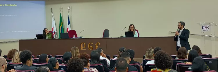 Auditório com várias pessoas sentadas em poltronas vermelhas, voltadas para uma mesa elevada onde duas mulheres estão sentadas com notebooks. À direita, um homem de terno fala em pé com microfone. Na parede ao fundo há bandeiras e um telão com apresentação projetada.