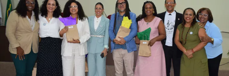 Oito pessoas posam sorridentes em frente ao palco de um auditório. O grupo está bem vestido, com roupas sociais e elegantes, e alguns seguram saquinhos de presente coloridos. Ao fundo, há bandeiras e um púlpito com o logotipo da PGE. O clima é de confraternização e encerramento de um evento.
