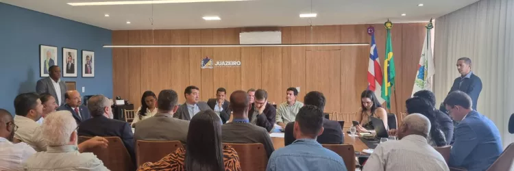 Sala de reuniões ampla e iluminada. Cerca de vinte pessoas adultas estão sentadas ao redor de uma mesa retangular, conversando e ouvindo. A maioria usa roupas sociais. Algumas pessoas anotam em cadernos ou usam notebooks e celulares. Ao fundo, há uma parede de madeira com um brasão e a palavra “Juazeiro”. À direita, três bandeiras em suportes verticais. À esquerda, quadros na parede azul. A cena mostra uma reunião formal em andamento.