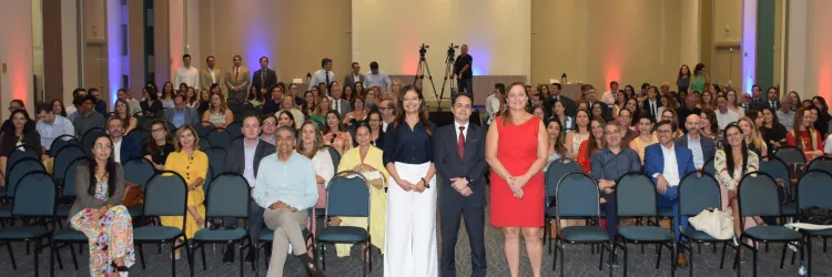 Foto geral do auditório com grande público e três pessoas em pé, ao centro, posando para a imagem. As cadeiras ocupam quase todo o espaço, mostrando alta participação. A estrutura de gravação e iluminação aparece ao fundo.