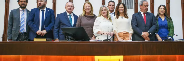 Imagem em ambiente institucional. Nove pessoas estão em pé, lado a lado, atrás de uma bancada de madeira. Elas usam roupas formais, como ternos, blazers, camisas e vestidos. A maioria olha para a frente e sorri. Sobre a bancada há microfones, papéis e um monitor. Ao centro do fundo, na parede, há um crucifixo. Abaixo da bancada, vê-se um painel com o logotipo e a sigla “ALBA”, da Assembleia Legislativa da Bahia. O local parece um plenário ou sala oficial.