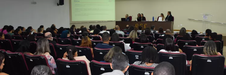 A imagem mostra um auditório cheio, com várias fileiras de cadeiras vermelhas ocupadas por pessoas adultas. Todos estão voltados para a frente, onde há uma mesa comprida com cinco pessoas sentadas, participando de uma apresentação. Um projetor exibe um slide com texto na parede. O ambiente é iluminado e organizado, com clima de palestra ou reunião formal. No canto direito, há um púlpito transparente com o logotipo “PGE”.