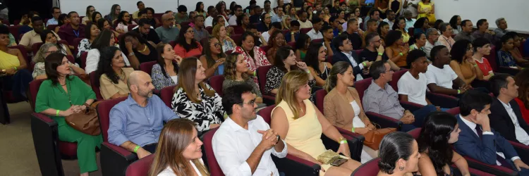 Auditório cheio, com muitas pessoas sentadas em poltronas vermelhas, assistindo ao evento