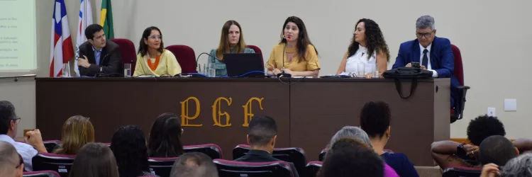 A imagem mostra seis pessoas sentadas lado a lado em uma mesa longa, participando de um evento formal. Há quatro mulheres e dois homens, todos vestidos de forma elegante. Atrás deles, estão três bandeiras: uma do Brasil, uma estadual e a terceira da PGE-BA. À frente da mesa, está a sigla "PGE" em letras grandes. Na parte inferior, algumas pessoas assistem à apresentação. O ambiente é uma sala de conferências com iluminação clara e um telão ao lado esquerdo. A cena transmite uma reunião oficial.
