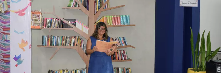 Uma mulher está em um espaço de leitura, em frente a uma estante em formato de árvore. Ela segura um livro aberto e sorri. O ambiente tem pufes coloridos, muitos livros, plantas e decoração com desenhos de pássaros. A cena transmite clima de leitura e evento cultural.