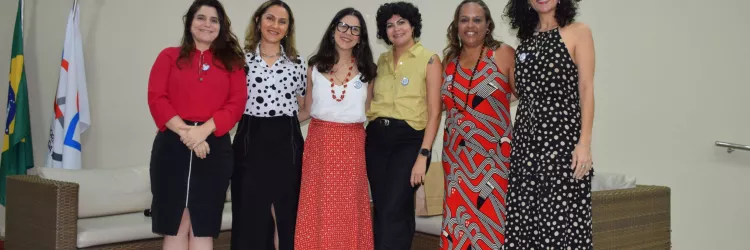 Seis mulheres posam sorrindo em frente aos sofás do auditório. Elas usam roupas coloridas e variadas, transmitindo diversidade. O ambiente é institucional e o grupo parece registrar o encerramento da atividade ou palestra