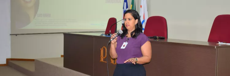Uma mulher fala ao microfone durante uma apresentação. Ela veste blusa roxa e crachá, posicionada diante de uma tela projetada. O ambiente parece ser uma sala de reunião ou auditório institucional.
