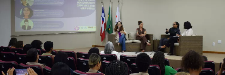 Em um auditório, várias pessoas estão sentadas em fileiras de cadeiras, formando o público e voltadas para o palco. No palco, quatro mulheres adultas estão sentadas em sofás claros, participando de uma roda de conversa. Uma delas segura o microfone e fala, enquanto as outras escutam. Ao fundo, um telão exibe informações do evento sobre fiscalização em tempo real e a Lei Maria da Penha. À direita do palco, estão as bandeiras da Procuradoria Geral, de Salvador e da Bahia. 