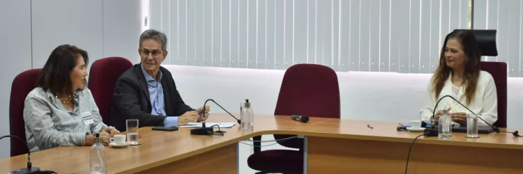 Imagem em ambiente interno, uma sala de reuniões. Três pessoas estão sentadas ao redor de uma mesa de madeira em formato angular. À esquerda, uma mulher de cabelo castanho curto usa roupa clara e conversa com um homem ao centro, que tem cabelos grisalhos, usa óculos e veste terno escuro com camisa clara. À direita, outra mulher, de cabelos longos e castanhos, veste blusa clara. Sobre a mesa há microfones, copos com água, xícaras de café e papéis. Ao fundo, parede clara com persianas verticais brancas.