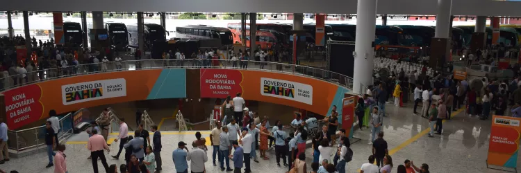 Vista ampla do interior da Nova Rodoviária da Bahia, em Salvador. O espaço é grande, com piso claro, teto alto e iluminação natural. Muitas pessoas circulam, conversam ou aguardam. Há escadas, filas organizadas e painéis com a identidade visual do Governo da Bahia. Ônibus estão visíveis ao fundo.
