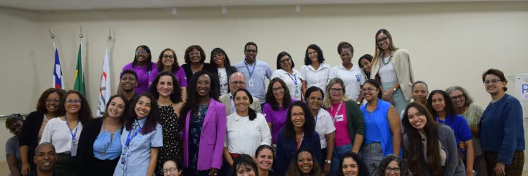 Grupo com cerca de 30 pessoas posa para foto em um auditório. A maioria é de mulheres, de diferentes idades e tons de pele, e há alguns homens. Estão organizadas em três filas, em pé e agachadas, todas sorrindo. Ao centro, uma mulher negra usa blazer roxo. Ao fundo, aparecem bandeiras e uma parede clara. No teto, há luzes embutidas e um projetor. Na frente, vêem-se cadeiras vermelhas. O ambiente é interno, iluminado e institucional.