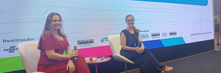 Em um palco, duas mulheres estão sentadas em poltronas brancas, lado a lado, cada uma segurando um microfone. À esquerda, uma mulher de vestido vermelho longo sorri para a câmera. À direita, outra mulher veste roupa escura e também sorri. Entre elas, há uma pequena mesa com objetos. Ao fundo, um telão grande exibe o título “Curso Introdutório – Marco Regulatório das Organizações da Sociedade Civil (MROSC)” e logotipos de instituições. O ambiente parece ser um evento ou palestra em Salvador.